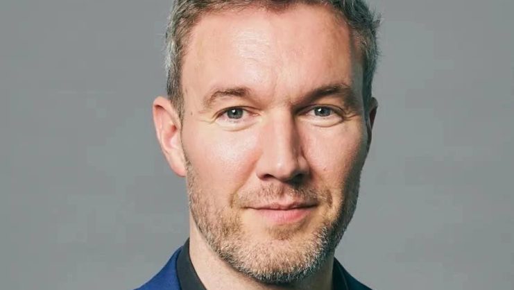 Erald Ghoos mit kurzen hellbraunen Haaren und gestutztem Bart trägt eine blaue Anzugsjacke und ein schwarzes Hemd und posiert vor einem schlichten grauen Hintergrund, der selbst auf einem Bärenmarkt das Vertrauen eines Bitcoin-Händlers erweckt.