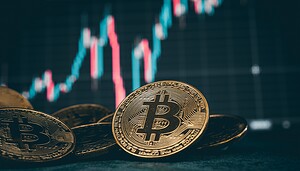 Mehrere physische Bitcoin-Münzen werden vor einem digitalen Bildschirm angezeigt, der ein schwankendes Bitcoin-Kursdiagramm mit roten und grünen Kerzen zeigt.
