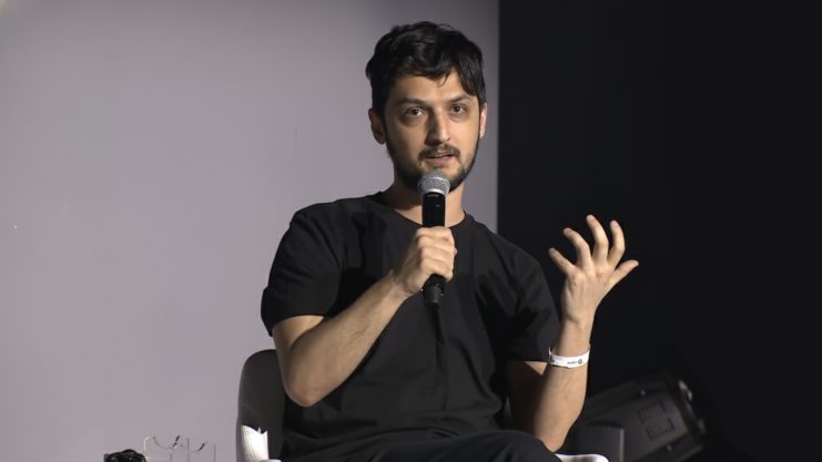 Eine Person in einem schwarzen Hemd sitzt auf einem Stuhl, hält ein Mikrofon in der Hand und gestikuliert mit der Hand, während sie über Krypto spricht.