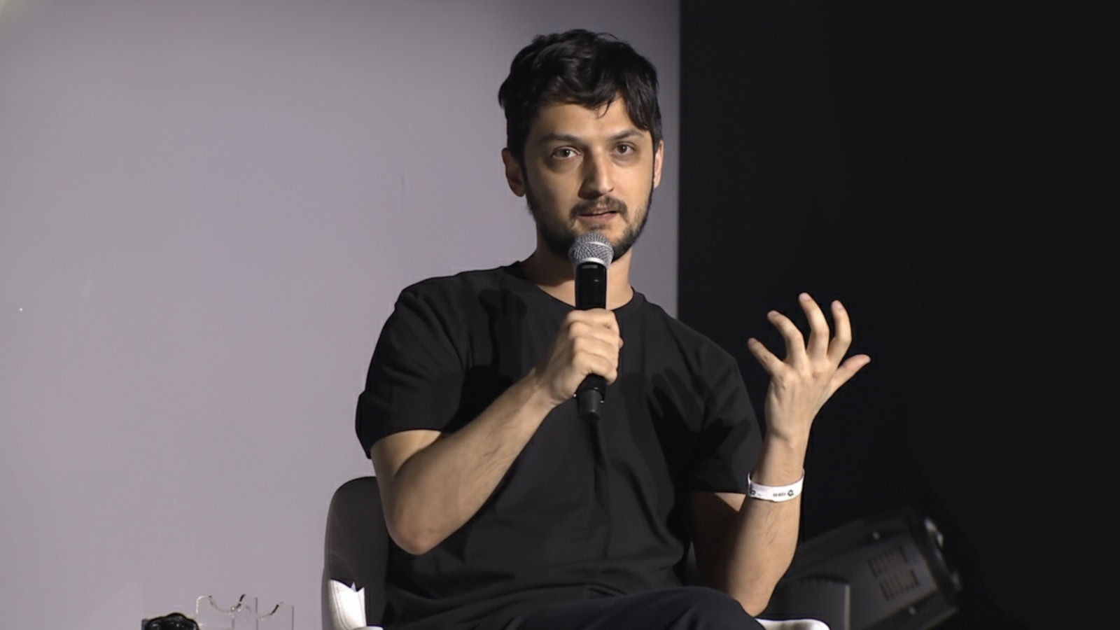 Eine Person in einem schwarzen Hemd sitzt auf einem Stuhl, hält ein Mikrofon in der Hand und gestikuliert mit der Hand, während sie über Krypto spricht.