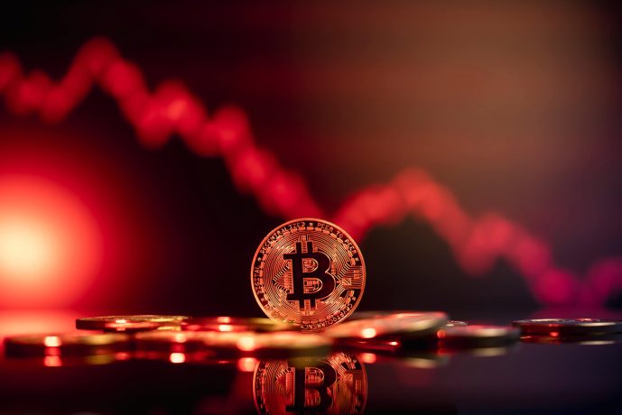 Eine Bitcoin-Münze steht aufrecht zwischen verstreuten Münzen mit einer roten, abwärts gerichteten Grafik im unscharfen Hintergrund, die den Rückgang des Kryptowertes hervorhebt.