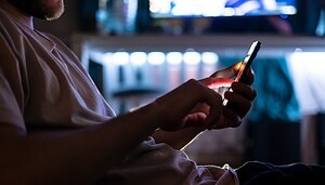 Eine Person in einem schwach beleuchteten Raum benutzt ein Smartphone, während im unscharfen Hintergrund ein Computermonitor und verschiedene Krypto-Objekte zu sehen sind.