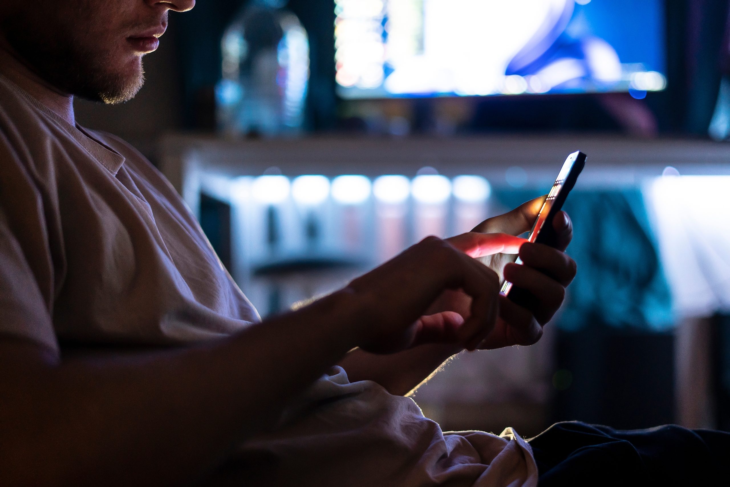 Eine Person in einem schwach beleuchteten Raum benutzt ein Smartphone, während im unscharfen Hintergrund ein Computermonitor und verschiedene Krypto-Objekte zu sehen sind.