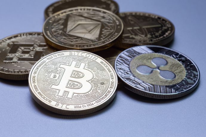 Mehrere physische Münzen, die verschiedene Kryptowährungen repräsentieren, darunter Bitcoin, Ethereum und Ripple, sind auf einer glatten, grauen Oberfläche angeordnet. Die herausragende Bitcoin-Münze sticht aus der Sammlung hervor.