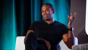 Krypto-Starinvestor Arthur Hayes gilt als erfahrener Makroexperte Arthur Hayes in einem schwarzen T-Shirt sitzt auf einem weißen Stuhl, gestikuliert mit einer Hand und spricht über Krypto vor einem dunklen, blaugrünen Hintergrund.