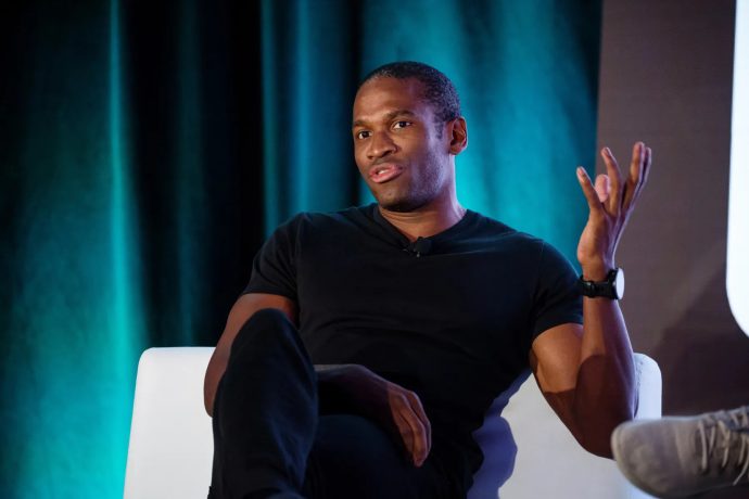 Arthur Hayes in einem schwarzen T-Shirt sitzt auf einem weißen Stuhl, gestikuliert mit einer Hand und spricht über Krypto vor einem dunklen, blaugrünen Hintergrund.