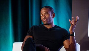 Arthur Hayes in einem schwarzen T-Shirt sitzt auf einem weißen Stuhl, gestikuliert mit einer Hand und spricht über Krypto vor einem dunklen, blaugrünen Hintergrund.