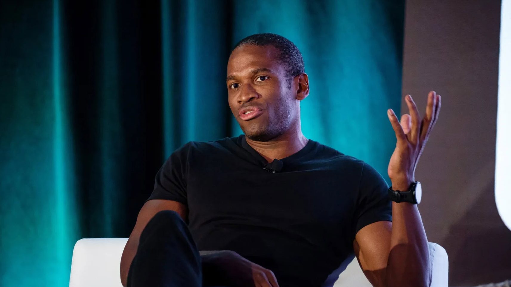 Arthur Hayes in einem schwarzen T-Shirt sitzt auf einem weißen Stuhl, gestikuliert mit einer Hand und spricht über Krypto vor einem dunklen, blaugrünen Hintergrund.