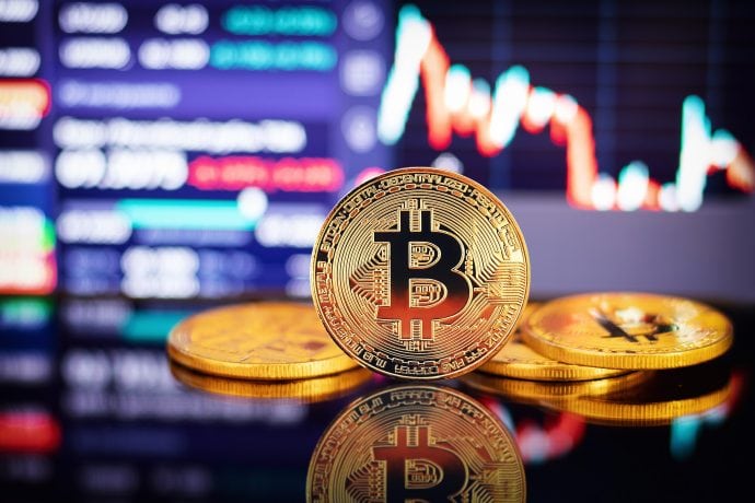 Eine physische Bitcoin-Münze wird vor einem verschwommenen Computerbildschirm angezeigt, der ein Finanzdiagramm mit roten und blauen Linien zeigt und so die dynamische Welt des Krypto-Handels einfängt.