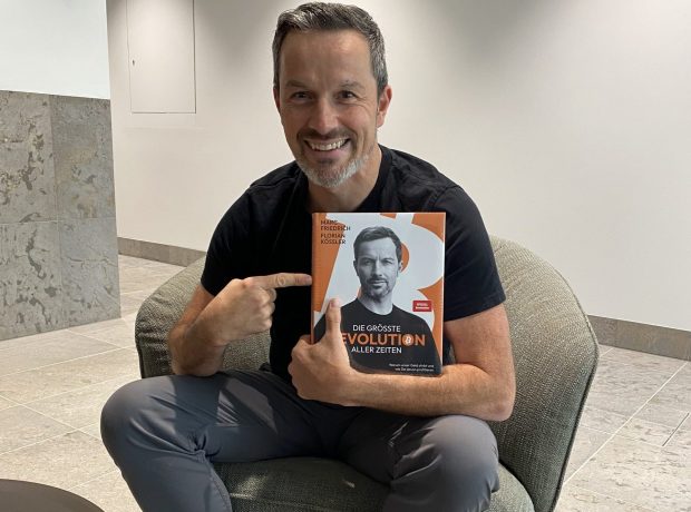 Der Finanzexperte Marc Friedrich präsentiert im Interview seinen aktuellen Bestseller „Die größte Revolution aller Zeiten“. In entspannter Atmosphäre, gekleidet in sportlich-bunten Sneakern, sitzt er auf einem Lounge-Sessel und hält das Buch mit einem breiten Lächeln in die Kamera. Das Cover zeigt ein Porträt von Friedrich – thematisch dreht sich das Werk um Systemwandel, wirtschaftliche Disruption und die Rolle von Bitcoin im Umbruch der Finanzwelt.