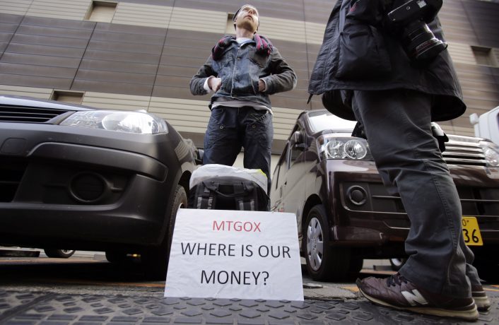 mt-gox-auszahlungen-verschoben Eine Person steht zwischen Autos mit einem Schild mit der Aufschrift "Mt.Gox WHERE IS OUR MONEY?" während einer scheinbaren Protestaktion oder Demonstration.