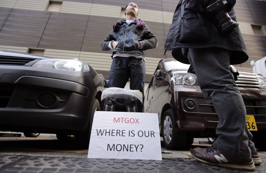 Eine Person steht zwischen Autos mit einem Schild mit der Aufschrift "Mt.Gox WHERE IS OUR MONEY?" während einer scheinbaren Protestaktion oder Demonstration.