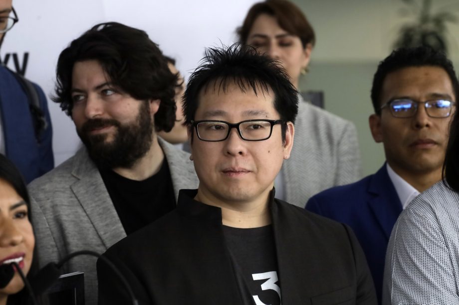 Eine Gruppe von Menschen in Geschäftskleidung steht zusammen, wobei ein Mann im Vordergrund eine Brille und ein schwarzes Jackett trägt und nach vorne blickt, während an seinem Revers eine dezente Bitcoin-Anstecknadel leuchtet.