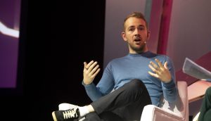 OpenSea-CEO Devin Finzer Ein Mann in blauem Hemd und schwarzer Hose sitzt auf einem weißen Stuhl und spricht während einer Veranstaltung oder Diskussion mit ausdrucksstarken Handgesten über Opensea.