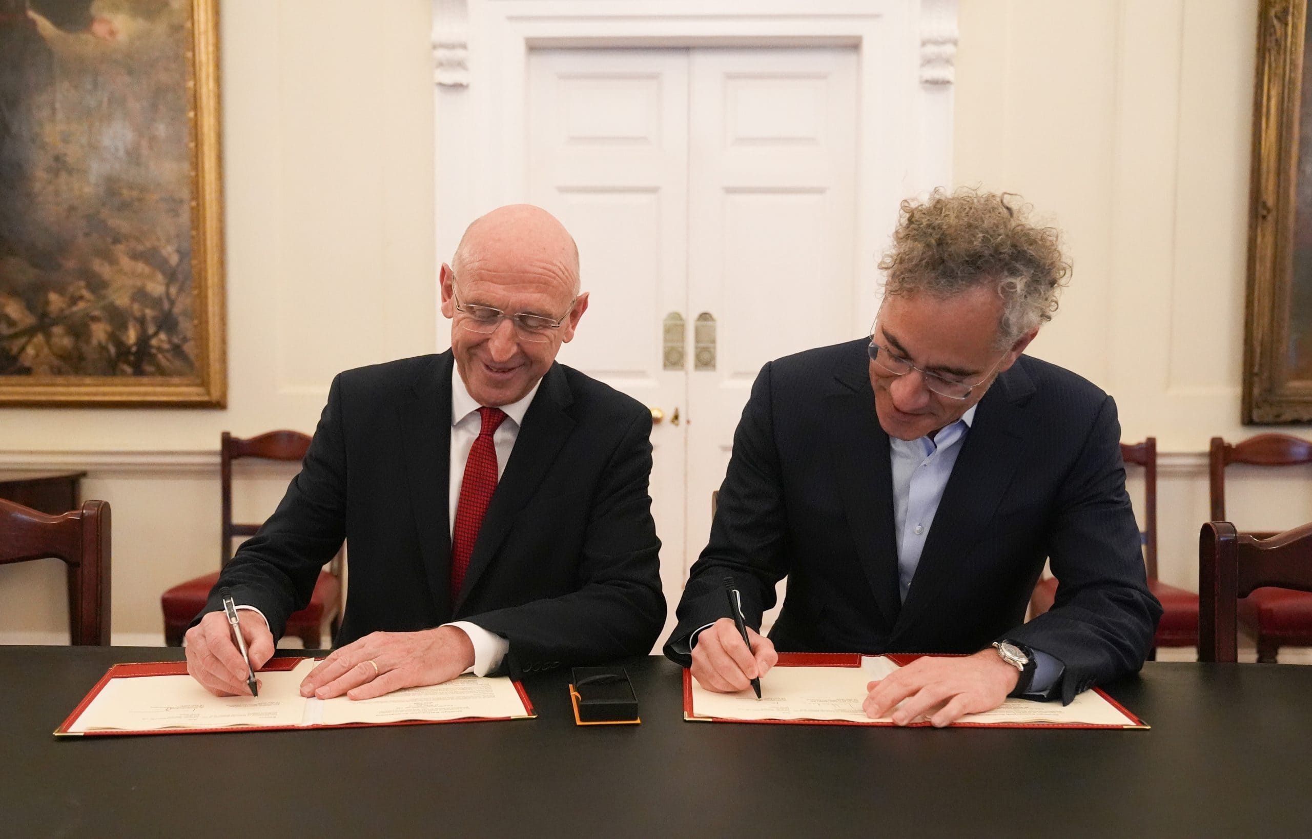 Zwei Männer in Anzügen sitzen an einem Tisch und unterschreiben Dokumente mit Palantir-Stiften in einem formellen Raum mit gerahmten Gemälden und weißen Doppeltüren im Hintergrund.