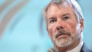 Ein Mann mittleren Alters mit grauem Haar und Bart spricht in ein Mikrofon über Bitcoin. Er trägt einen Anzug und eine orangefarbene Krawatte vor einem verschwommenen hellblauen Hintergrund.