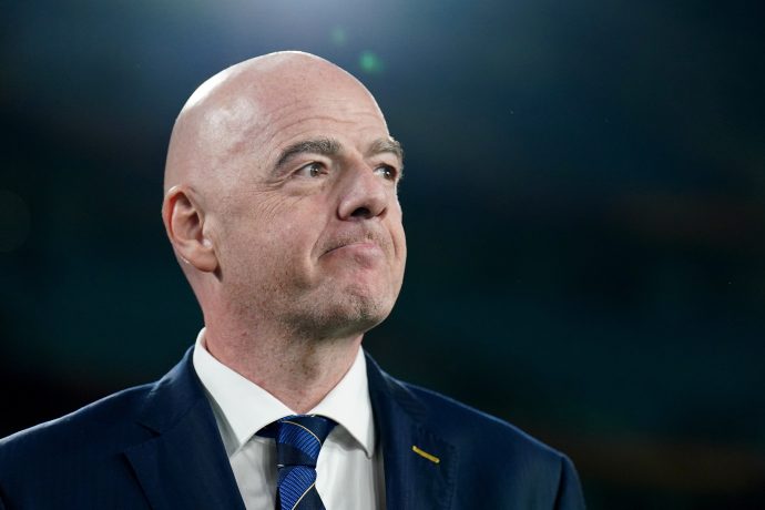 Ein glatzköpfiger Mann in Anzug und gestreifter Krawatte blickt mit neutraler Miene zur Seite und steht vor einem dunklen, unscharfen Hintergrund - vielleicht bei einer Veranstaltung im Zusammenhang mit der FIFA-NFT-Plattform oder der Schweizer Glücksspielaufsicht.