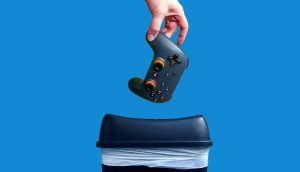 Der Blockchain-Gaming-Markt hinkt seinen Erwartungen hinterher Eine Hand hält einen schwarzen Videospiel-Controller über einem Mülleimer vor einem blauen Hintergrund.