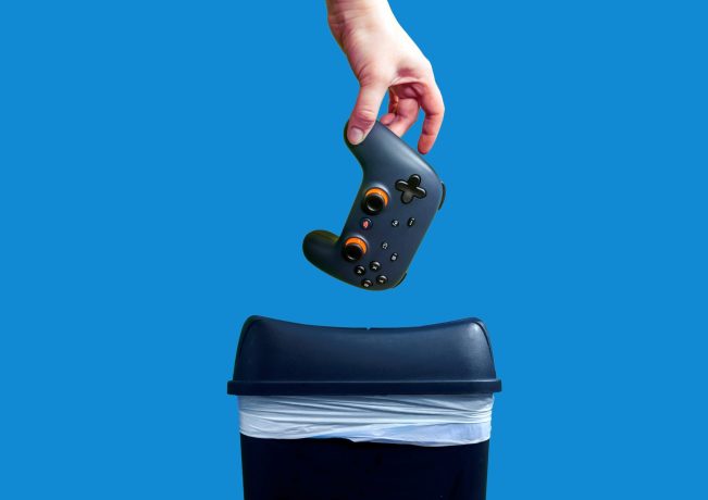 Eine Hand hält einen schwarzen Videospiel-Controller über einem Mülleimer vor einem blauen Hintergrund.