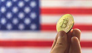 Eine Hand hält eine goldfarbene Bitcoin-Münze vor einer verschwommenen amerikanischen Flagge, die die wachsende Präsenz von Bitcoin in den Vereinigten Staaten symbolisiert.