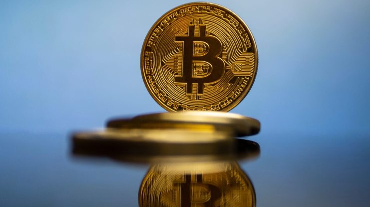 Bitcoins,Mirroring,Each,Other,Against,A,Blue,Background Eine goldene Bitcoin-Münze steht aufrecht, ihr Spiegelbild ist auf einer glänzenden Oberfläche sichtbar und symbolisiert Stärke im Krypto-Markt, während andere Münzen flach im Hintergrund liegen.