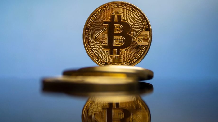 Eine goldene Bitcoin-Münze steht aufrecht, ihr Spiegelbild ist auf einer glänzenden Oberfläche sichtbar und symbolisiert Stärke im Krypto-Markt, während andere Münzen flach im Hintergrund liegen.