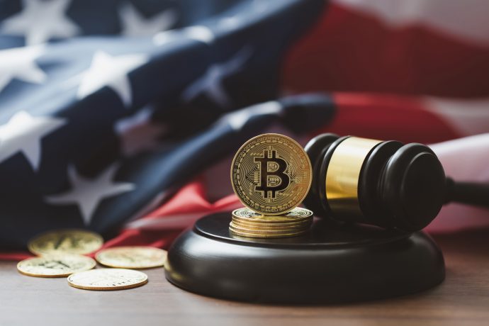 Eine Bitcoin-Münze, die den Einfluss von Figuren wie dem Bitcoin-Jesus Roger Ver symbolisiert, ruht auf dem Hammer eines Richters mit anderen Münzen in der Nähe, vor einem verschwommenen Hintergrund mit der Flagge der Vereinigten Staaten.