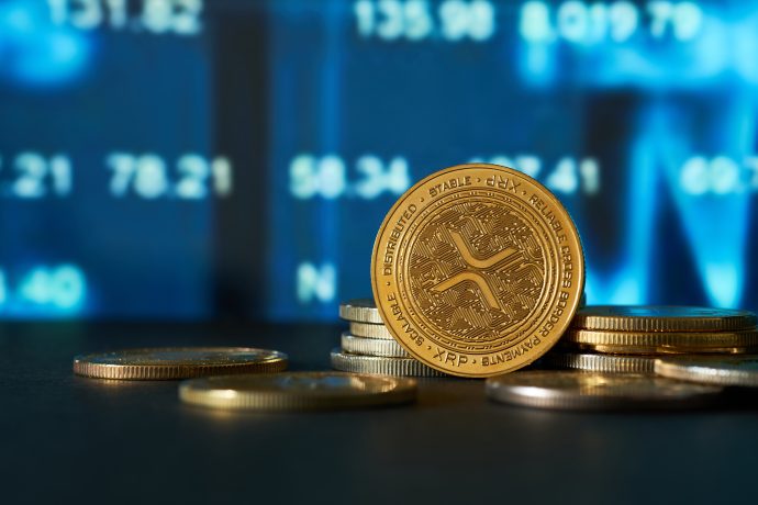 Ein Stapel Ripple-Münzen (XRP) wird vor einem digitalen Bildschirm gezeigt, der Finanzdaten und Börsenzahlen anzeigt.