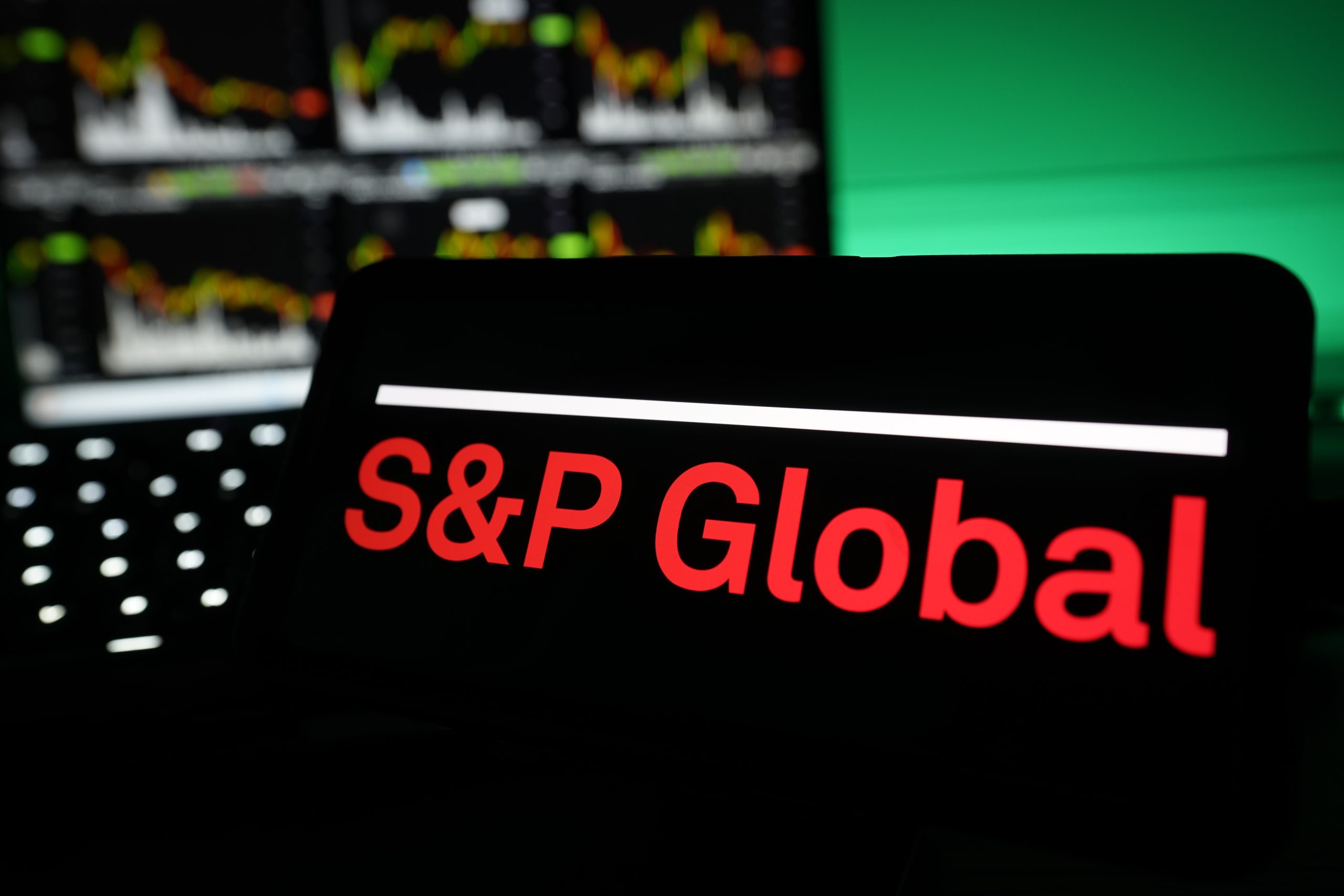 Auf einem Bildschirm wird das Logo von S&P Global in roter Schrift angezeigt, während im Hintergrund unscharfe S&P-Finanzcharts und -Grafiken zu sehen sind.
