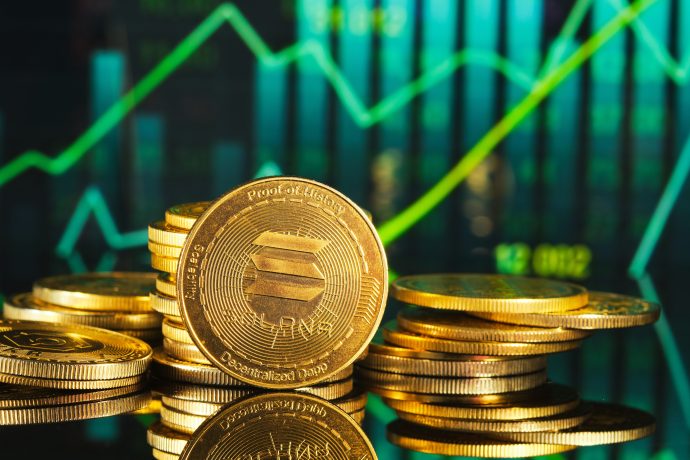 Stapel von Goldmünzen im Proof-of-Stake-Design und mit Solana-Branding liegen vor einem Bildschirm, der steigende Finanzcharts und grüne Aufwärtstrends anzeigt.