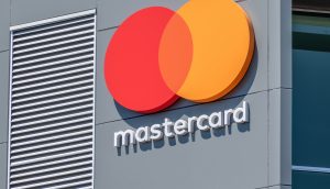 Das Mastercard-Logo mit sich überschneidenden roten und gelben Kreisen und weißem Kleinbuchstaben ist auf einer grauen Gebäudehülle abgebildet und spiegelt die wachsende Präsenz von Mastercard im Bereich der digitalen Zahlungen und Stablecoins wider.