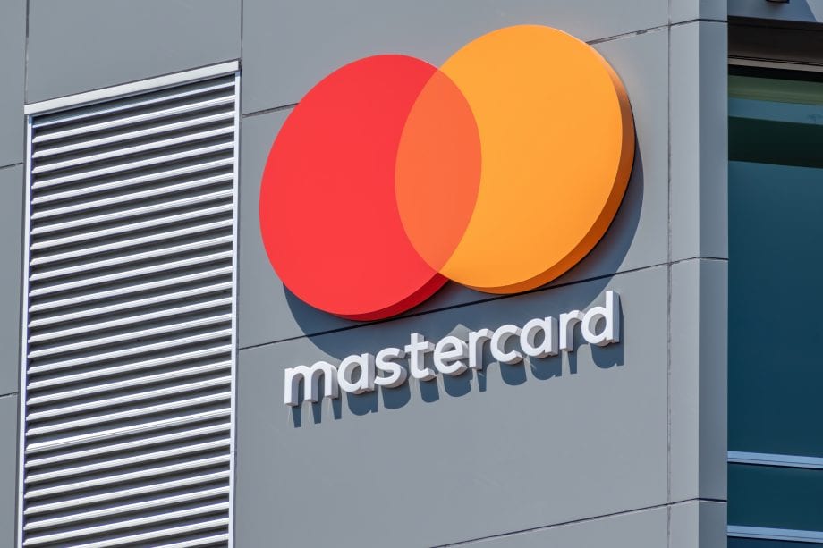 Das Mastercard-Logo mit sich überschneidenden roten und gelben Kreisen und weißem Kleinbuchstaben ist auf einer grauen Gebäudehülle abgebildet und spiegelt die wachsende Präsenz von Mastercard im Bereich der digitalen Zahlungen und Stablecoins wider.
