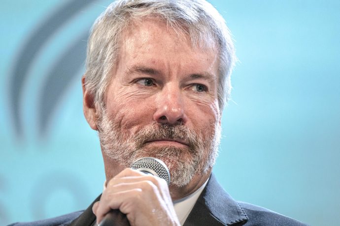 Ein Mann mit grauem Haar und Bart, der einen Anzug trägt, hält ein Mikrofon in der Hand und blickt vor einem unscharfen blauen Hintergrund zur Seite, während er scheinbar tief in Gedanken eine Strategie diskutiert.