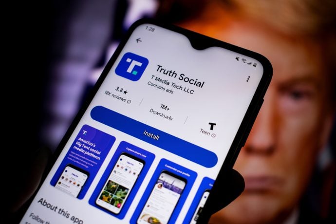 trump-truth-social-krypto-wetten Auf einem Smartphone-Bildschirm wird die Installationsseite der Truth Social-App auf Google Play angezeigt, wobei ein Teil des Gesichts einer Person im Hintergrund unscharf dargestellt ist, was Trumps Social-Media-Plattform hervorhebt.