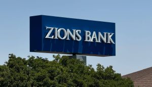 Die US-Bank Zions steht derzeit im Fokus des Finanzmarkts Auf einem blauen rechteckigen Schild mit weißen Buchstaben steht ZIONS BANK, ein Name, der im Zusammenhang mit der US-Bankenkrise und Diskussionen über Bitcoin häufig genannt wird. Das Schild ist über grünen Bäumen mit einem klaren Himmel im Hintergrund angebracht.