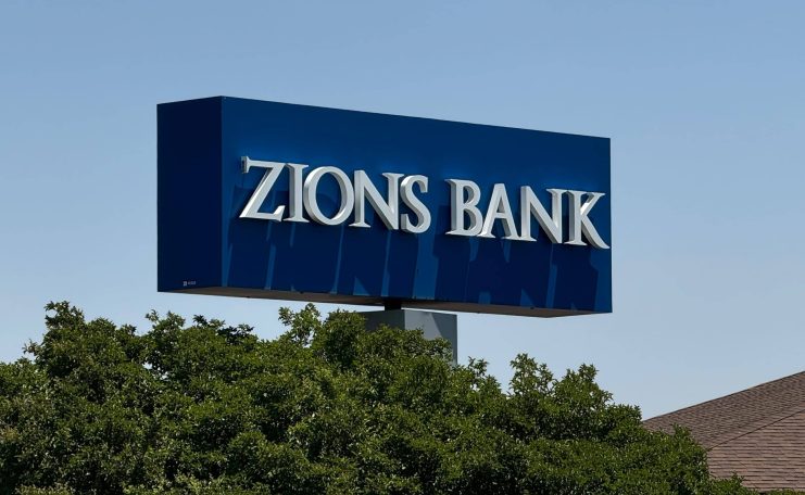Auf einem blauen rechteckigen Schild mit weißen Buchstaben steht ZIONS BANK, ein Name, der im Zusammenhang mit der US-Bankenkrise und Diskussionen über Bitcoin häufig genannt wird. Das Schild ist über grünen Bäumen mit einem klaren Himmel im Hintergrund angebracht.