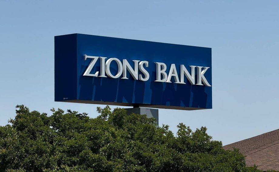 Auf einem blauen rechteckigen Schild mit weißen Buchstaben steht ZIONS BANK, ein Name, der im Zusammenhang mit der US-Bankenkrise und Diskussionen über Bitcoin häufig genannt wird. Das Schild ist über grünen Bäumen mit einem klaren Himmel im Hintergrund angebracht.