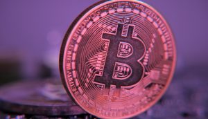 Eine Nahaufnahme einer physischen Bitcoin-Münze, die aufrecht vor einem unscharfen Hintergrund steht und mit lila und rosa Licht beleuchtet wird - ein Bild, das an einen Bitcoin-Wal erinnert, der die Krypto-Szene dominiert.