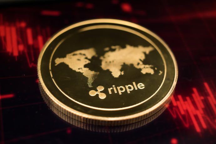 Eine goldene XRP-Münze mit dem Ripple-Logo und einer Weltkarte ruht auf einer dunklen Oberfläche, die von roten Finanzdiagrammlinien beleuchtet wird.