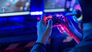 Ein Gamer trägt ein Headset und hält einen Controller in der Hand, während er vor zwei Monitoren in einem schwach beleuchteten, blau gestalteten Raum eine Spielesitzung abhält.