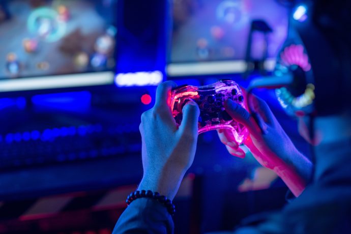 Warum Blockchain-Games dem Hype nie gerecht werden Ein Gamer trägt ein Headset und hält einen Controller in der Hand, während er vor zwei Monitoren in einem schwach beleuchteten, blau gestalteten Raum eine Spielesitzung abhält.