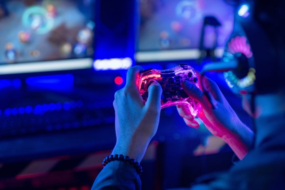 Ein Gamer trägt ein Headset und hält einen Controller in der Hand, während er vor zwei Monitoren in einem schwach beleuchteten, blau gestalteten Raum eine Spielesitzung abhält.