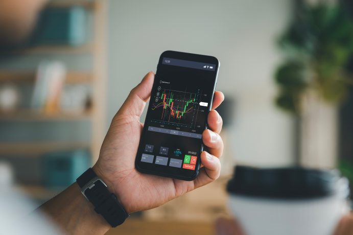 Eine Person hält ein Smartphone, auf dem ein Aktienchart angezeigt wird, während sie mit der anderen Hand einen Einweg-Kaffeebecher umklammert.
