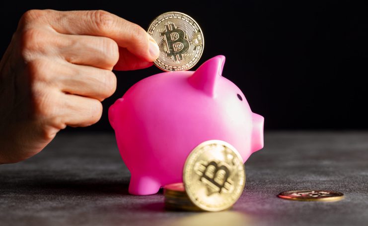 Eine Hand legt eine Bitcoin-Münze in ein rosafarbenes Sparschwein, auf dem weitere Bitcoin-Münzen gestapelt und auf dem Tisch verstreut sind, die das Bitcoin-Sparen symbolisieren.