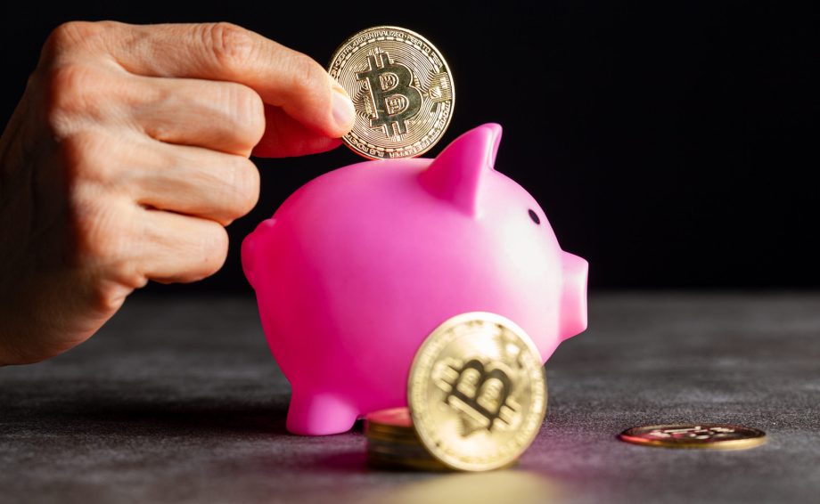 Eine Hand legt eine Bitcoin-Münze in ein rosafarbenes Sparschwein, auf dem weitere Bitcoin-Münzen gestapelt und auf dem Tisch verstreut sind, die das Bitcoin-Sparen symbolisieren.