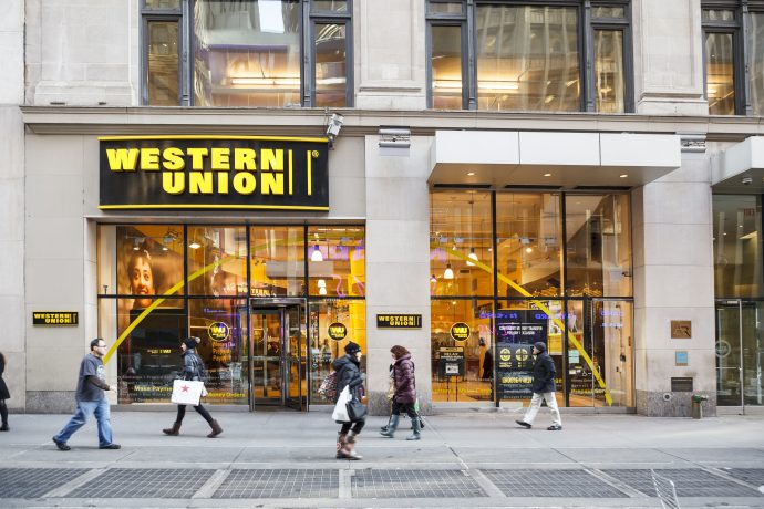 western-union-krypto-stablecoin Fußgänger gehen an einer Western-Union-Filiale mit großen gelben Schildern in einer Stadtstraße vorbei, wobei die Glasfenster der Schaufensterfront das Innere der Western Union hervorheben.