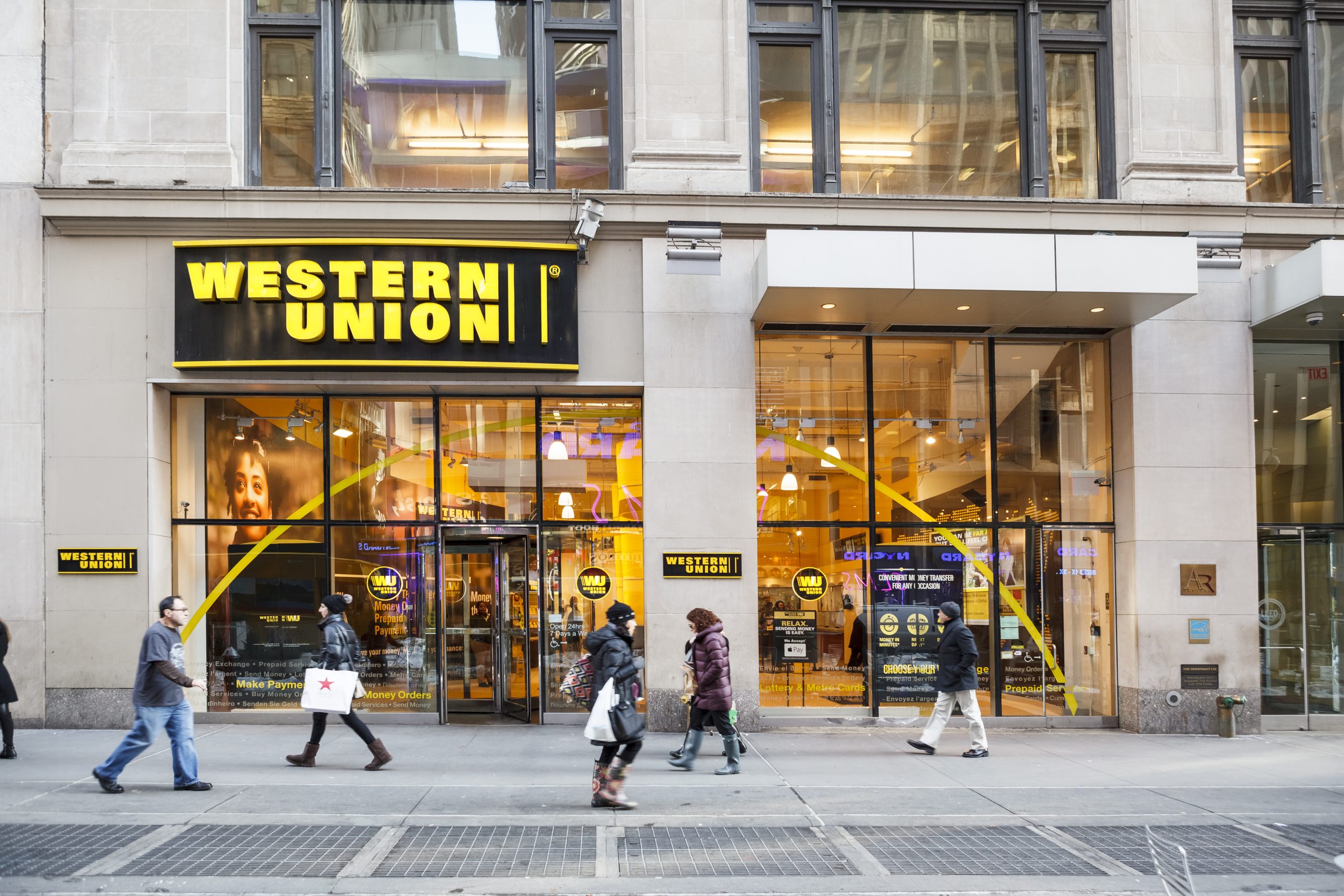 western-union-krypto-stablecoin Fußgänger gehen an einer Western-Union-Filiale mit großen gelben Schildern in einer Stadtstraße vorbei, wobei die Glasfenster der Schaufensterfront das Innere der Western Union hervorheben.