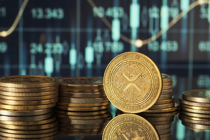 Stapel von goldenen XRP-Kryptowährungsmünzen sind auf einer spiegelnden Oberfläche angeordnet, mit einem XRP-Börsen-Candlestick-Chart im unscharfen Hintergrund.