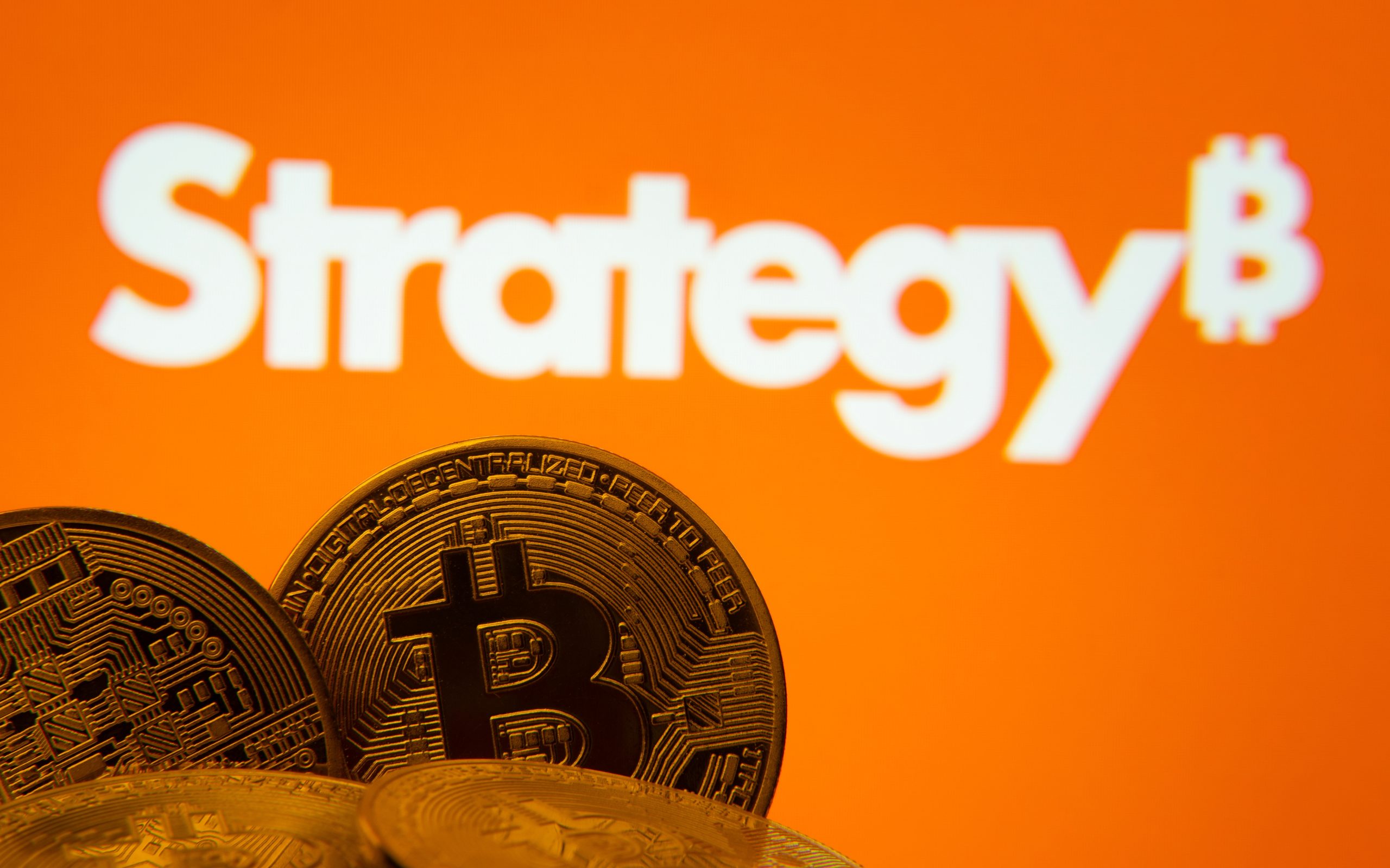 Mehrere physische Bitcoin-Münzen vor einem orangefarbenen Hintergrund mit dem Wort "Strategy" und einem markanten Bitcoin-Symbol.