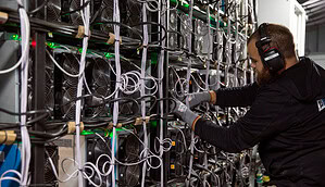 Ein Mann mit Kopfhörern und Handschuhen arbeitet an einer großen Reihe von Bitcoin-Mining-Maschinen mit zahlreichen Kabeln in einer industriellen Umgebung.